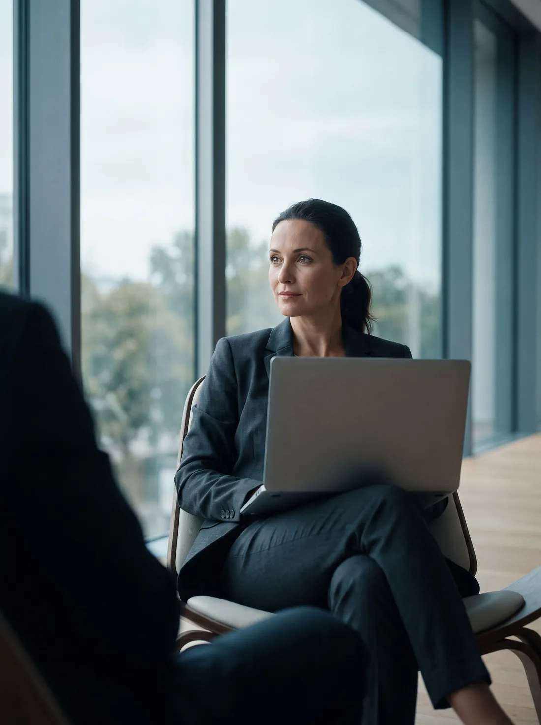 An accountant in a charcoal suit seated by a tall window with a laptop, turning toward a client mid-conversation.